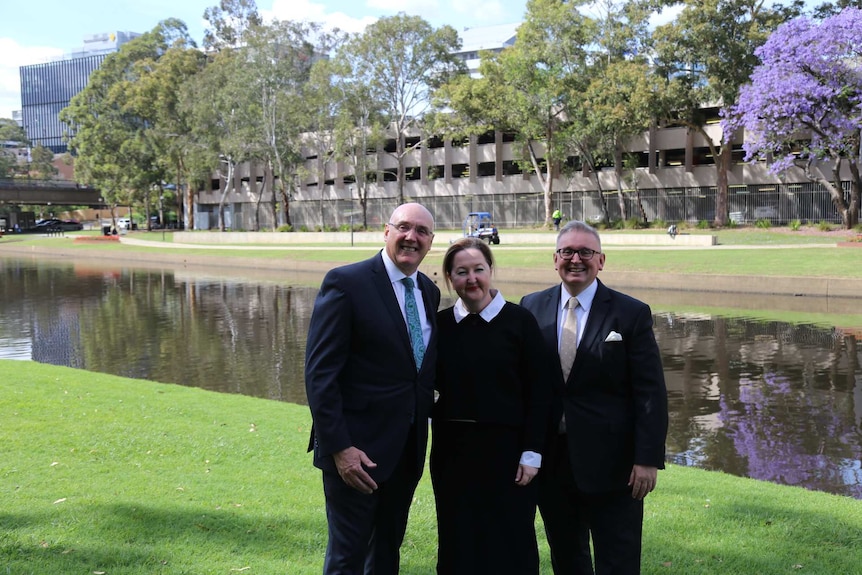 Women in centre, flanked by men in suits on either side, standing on grass by riverbank, sunshine.