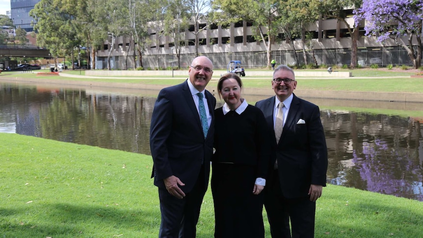 Women in centre, flanked by men in suits on either side, standing on grass by riverbank, sunshine.