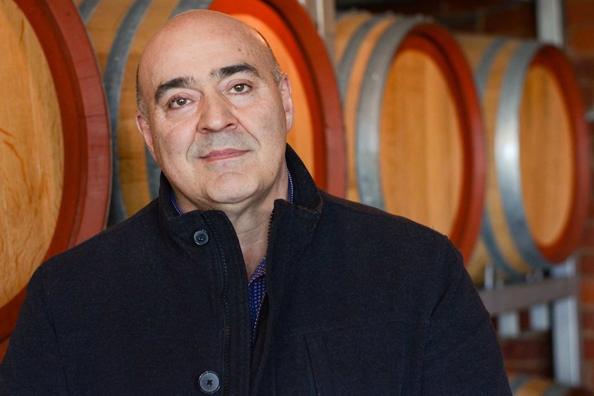 A man in a black coat with wine barrels behind him.