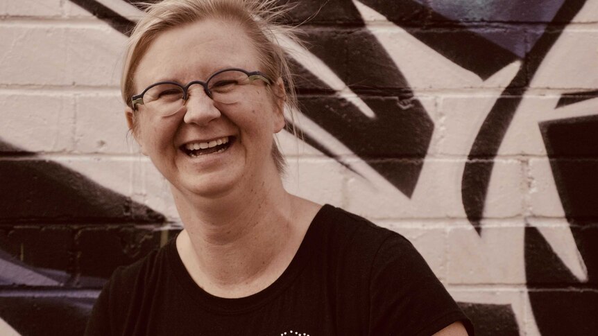 Image of a smiling woman wearing glasses in front of a graffiti-covered wall.