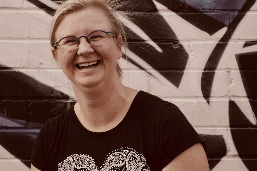 Image of a smiling woman wearing glasses in front of a graffiti-covered wall.