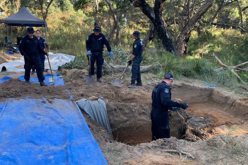 A police officer stands in a knee deep hole while three officers look on while holding shovels.