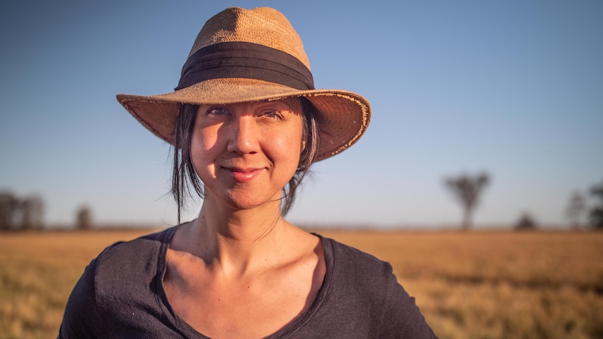 A woman wearing a hat stands in a golden paddock of wheat, stares at the camera.