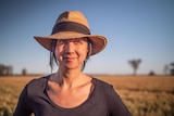 A woman wearing a hat stands in a golden paddock of wheat, stares at the camera.