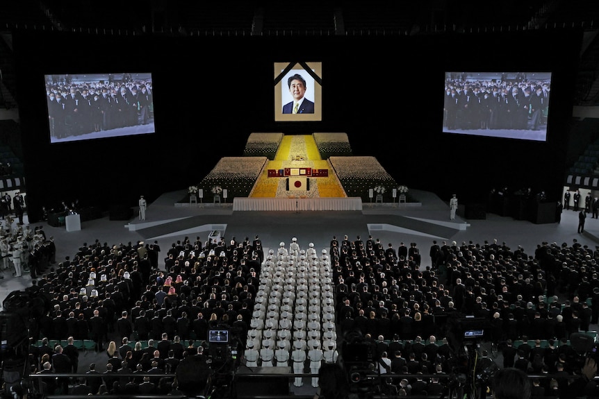 A large crowd of people in dark formal dress sit before a large stage set up for a state funeral.