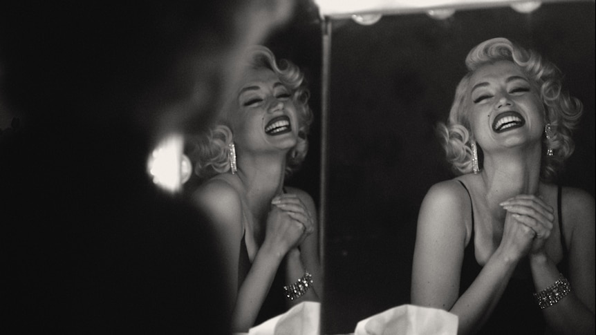 Black and white image of striking white woman with curly 50s style blonde hair beaming at her reflection in dressing room mirror