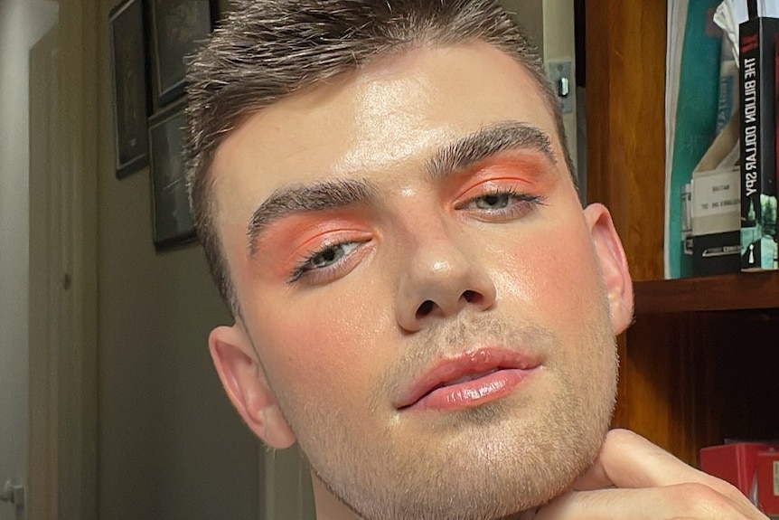 Davis Atkin in rose eye make-up posing for a selfie with one hand to his head, which is tilted.