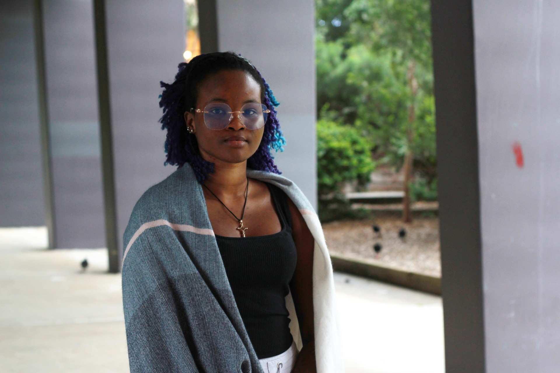 A young woman wears a shawl over her shoulders standing under a covered exterior walkway on a grey day.