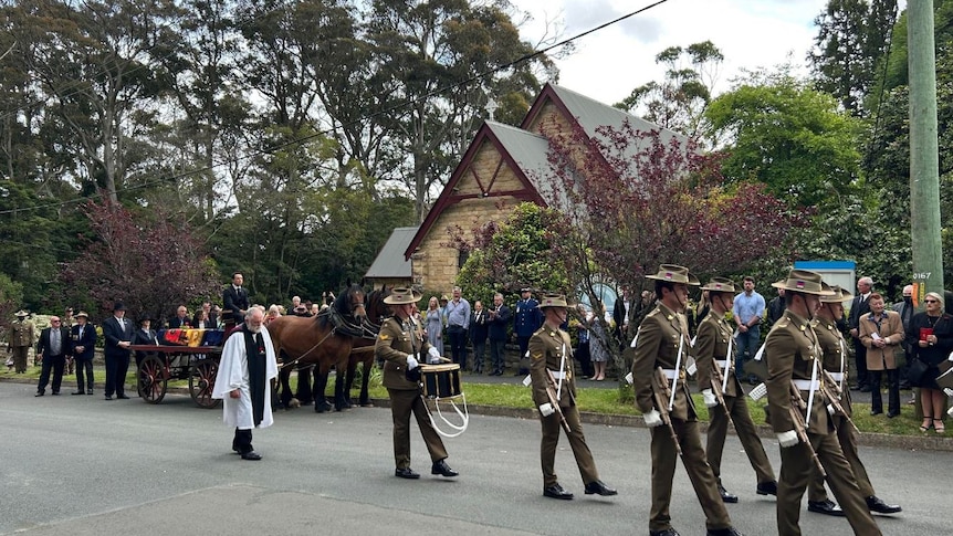 Marching soldiers lead a horse drawn funeral procession outside a sandstone church.