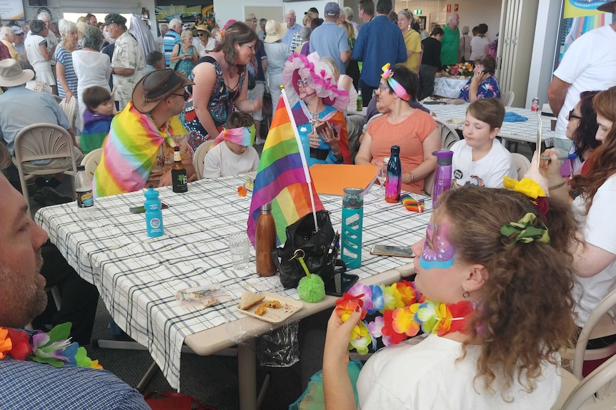 a group of people sit at a table wearing rainbow colours with a rainbow flag on the table