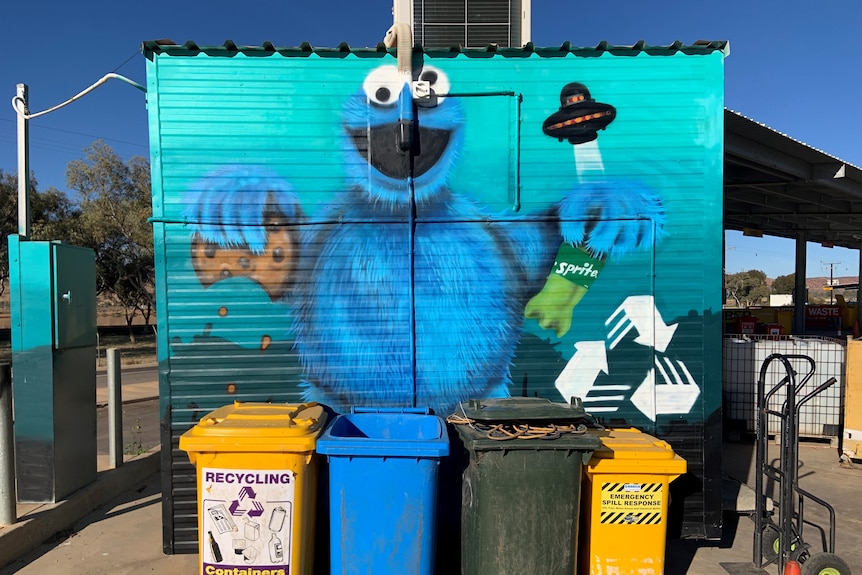 Wall mural of Cookie Monster holding chocolate chip cookie and sprite bottle. Four bins in front of the wall. 