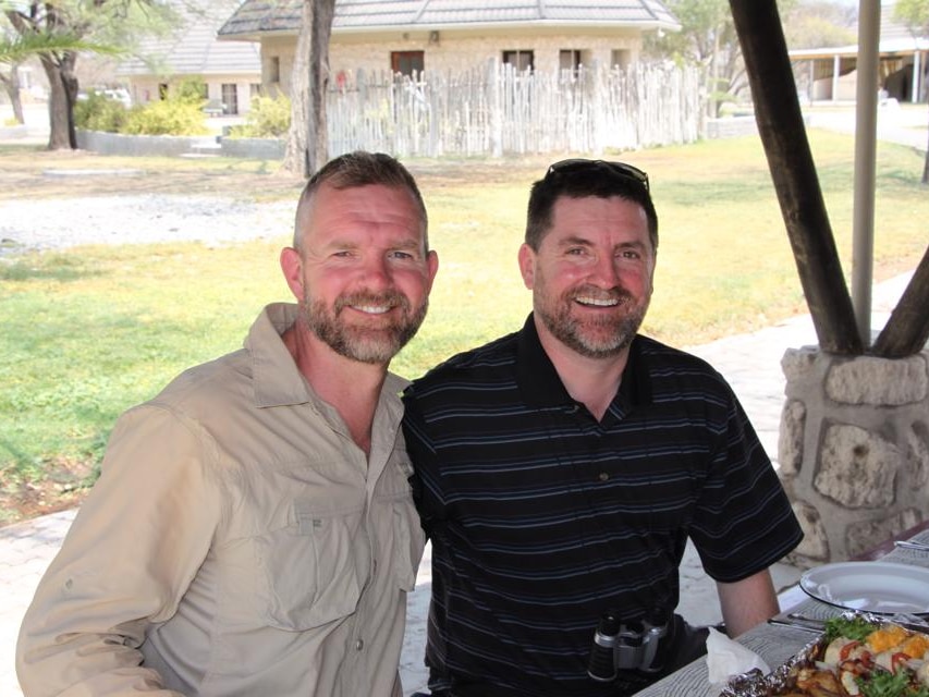 Two men smiling and sitting at an undercover picnic area.