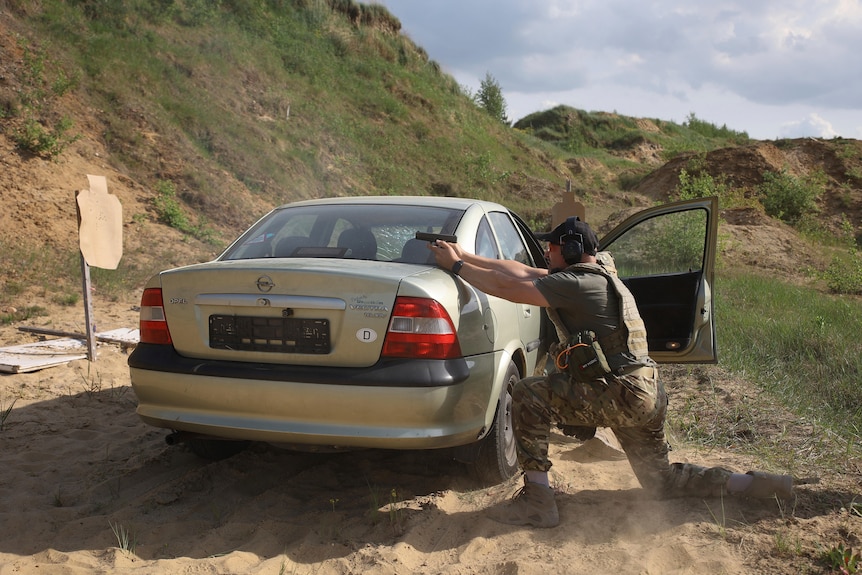 A man in fatigues uses a small grey car for cover as he practices shooting a pistol held with two hands over the car's boot. 