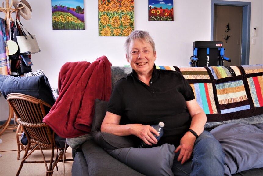 An older woman with short, grey hair sits on  a lounge chair, wearing a black polo shirt
