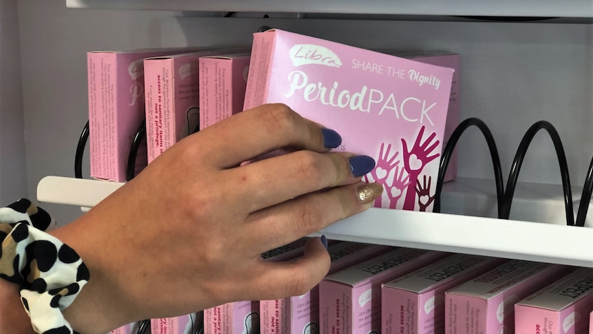 A hand places a small pink box containing sanitary items into a vending machine.