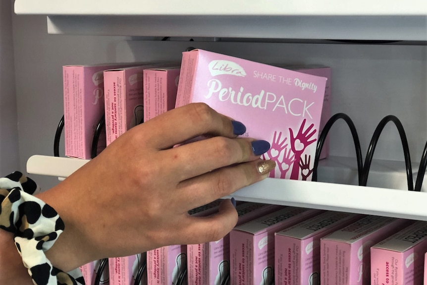 A hand places a small pink box containing sanitary items into a vending machine.