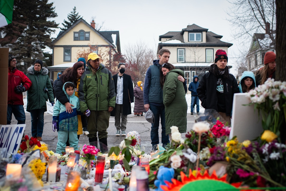 Adults and children look at a memorial on a street featuring flowers and candles.