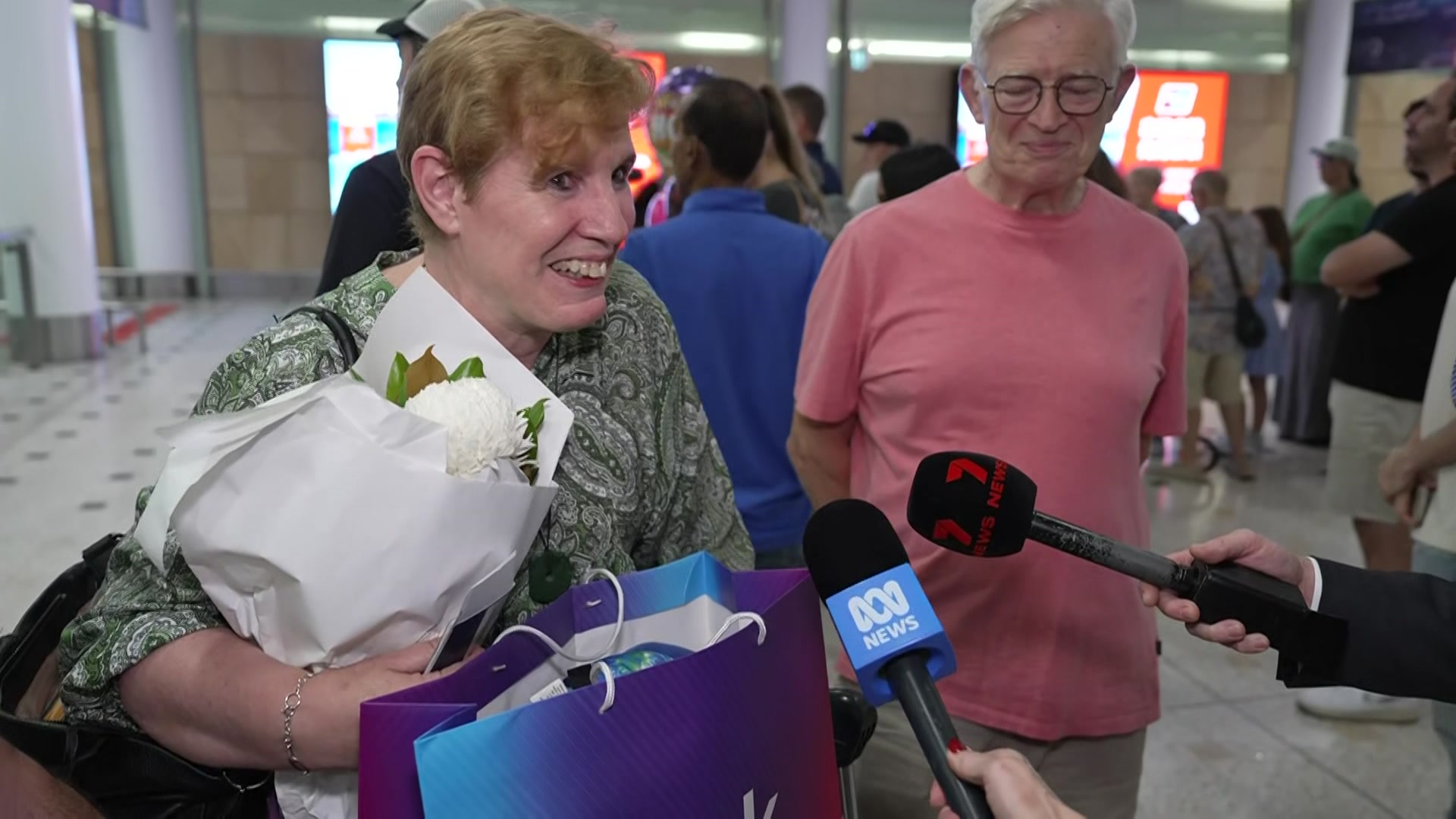 Una mujer de mediana edad sostiene flores mientras sonríe y habla con los periodistas en un aeropuerto.