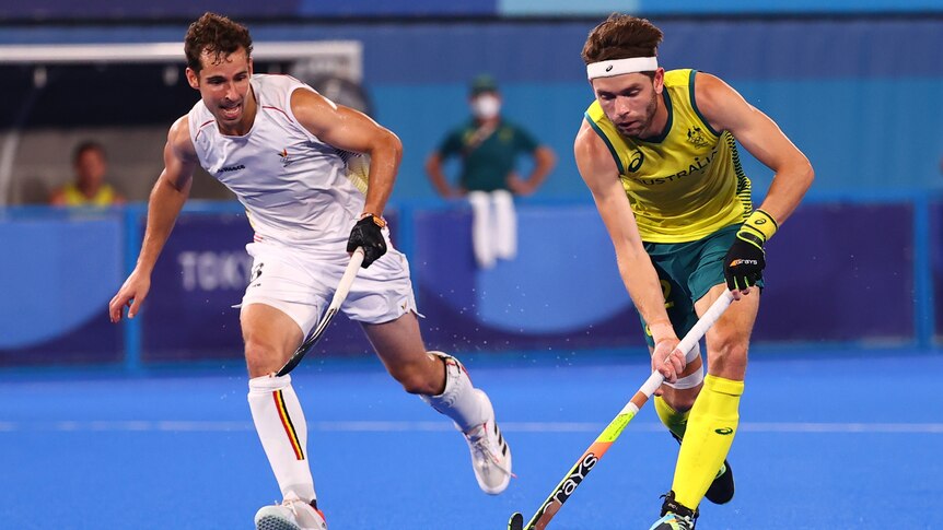 An Australian male hockey player controls the ball with his stick as a Belgian opponent runs alongside him.