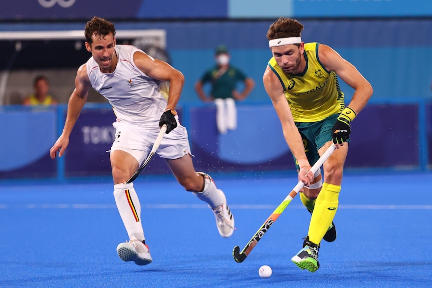 An Australian male hockey player controls the ball with his stick as a Belgian opponent runs alongside him.