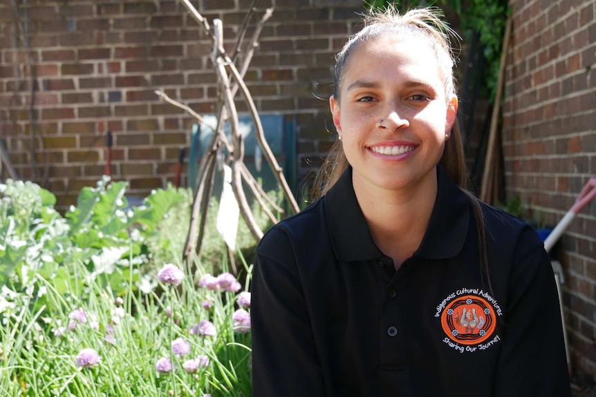 A woman wearing a black polo shirt is sitting in a garden surrounded by a plant with purple flowers, smiling at the camera.