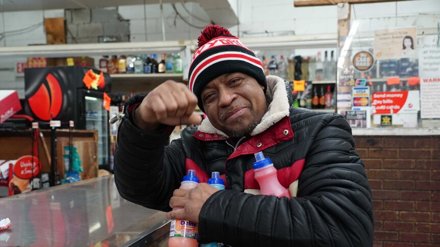 Duke God holds a couple of drink bottles and raises his fist in the air in a US shop