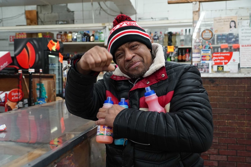 Duke God holds a couple of drink bottles and raises his fist in the air in a US shop