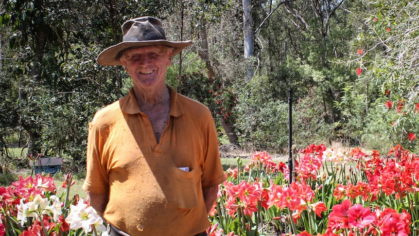 An old man wearing a hat and a grin stands in front of his garden of beautiful red flowers