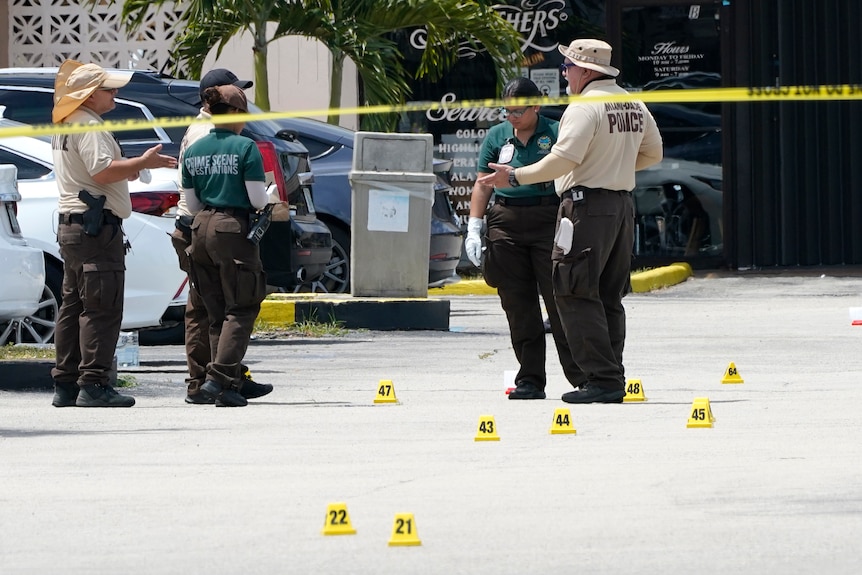 Five people in police and crime scene uniforms gather around numbered pieces of evidence on the ground. 