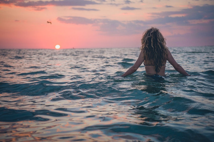 A photo of a woman in beach water facing the sunset with wavy blonde and brown hair