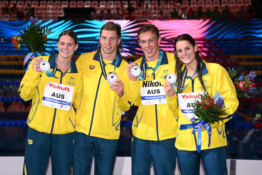 Four Australian swimmers, two female, two male, stand smiling on the podium with silver medals.