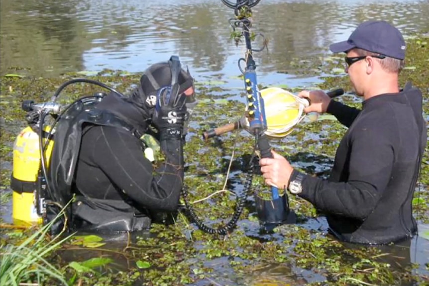 Two police divers wading in a pond with one holding a metal detector and one listening on headphones.