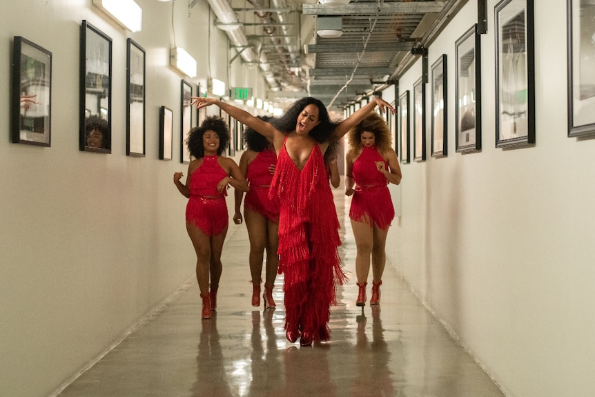 A woman in red flapper dress with arms raised in joy leads four others down long hallway lined with B&W framed pictures.