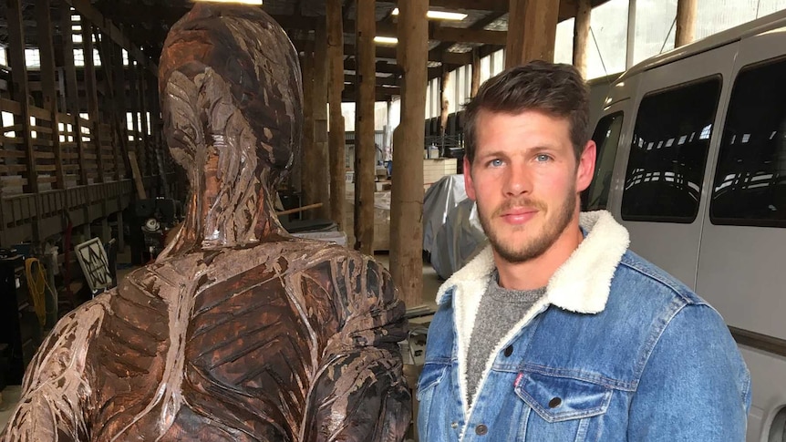 Adam Humphries is standing beside a wooden figure he's carved from a single piece of wood.