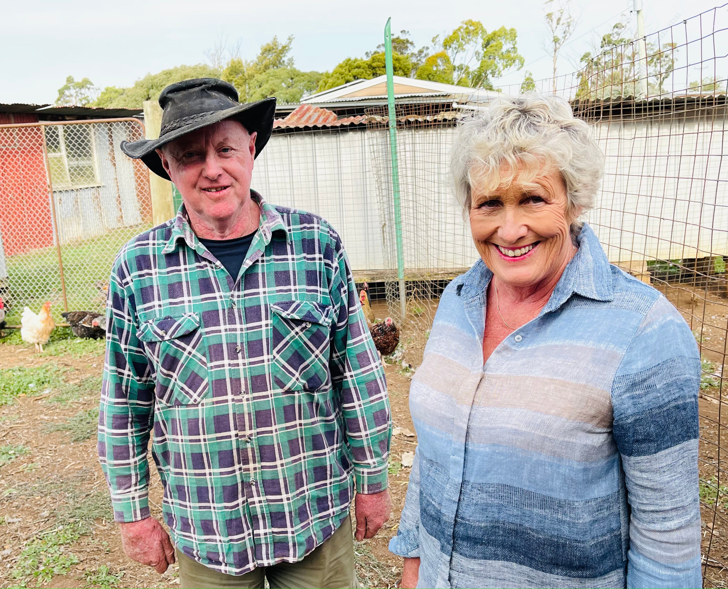 Man and woman smiling and standing outdoors in a chicken enclosure. 
