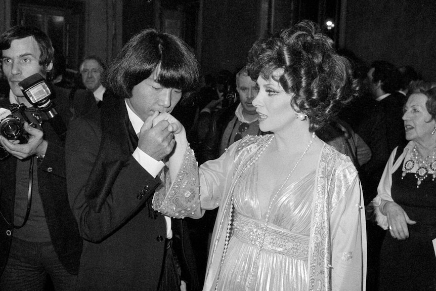 You see a black and white photo of a young Kenzo Takada kiss the hand of Gina Lollobrigida in a Grecian-style dress.