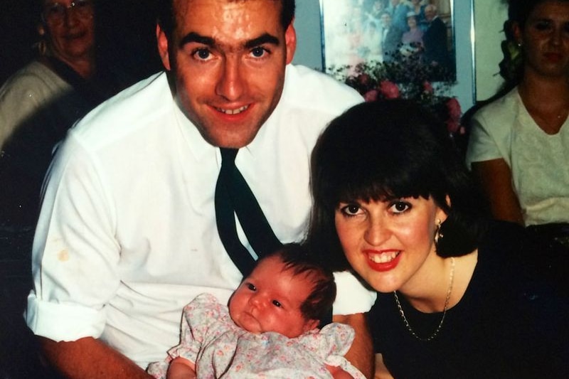 A man and a woman in formal clothing, smiling, hold a small baby girl, wearing a dress.