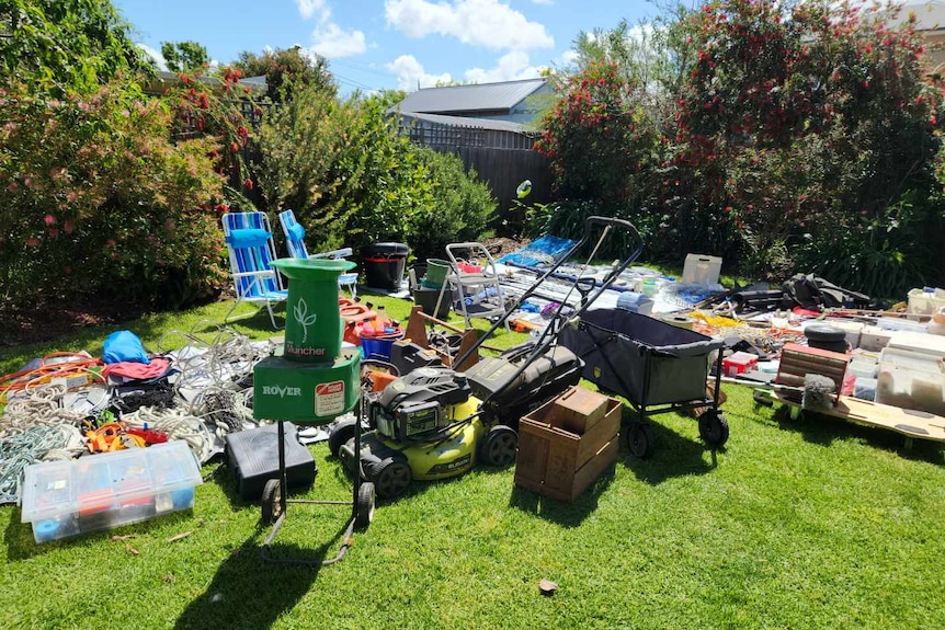 Many items for sale are spread across a lawn, including a lawnmower, mulcher, deck chairs and power cords.