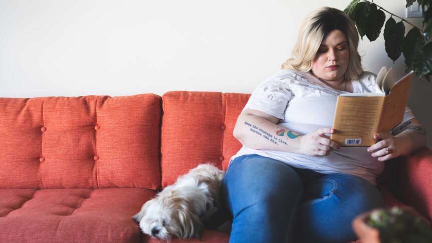 a pretty blonde woman sits on a red couch with her dog.  she's reading a book you might notice she's overweight but you may not