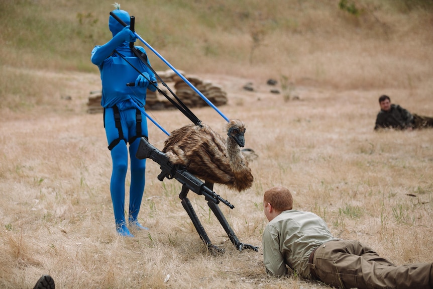 A person in a blue morph suit controls an emu puppet pointing a gun at a solider in a field