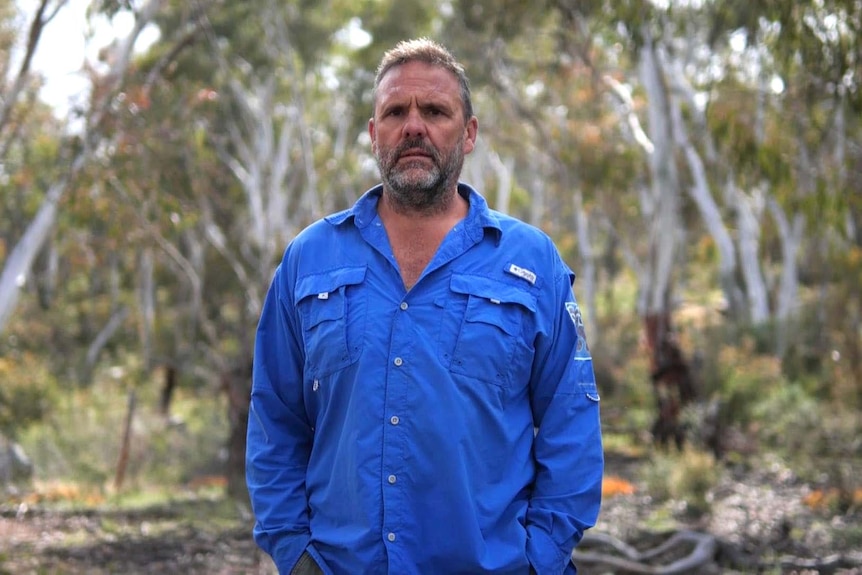 A man stands, his hands in his pockets and a blank expression. He is standing in the bush.