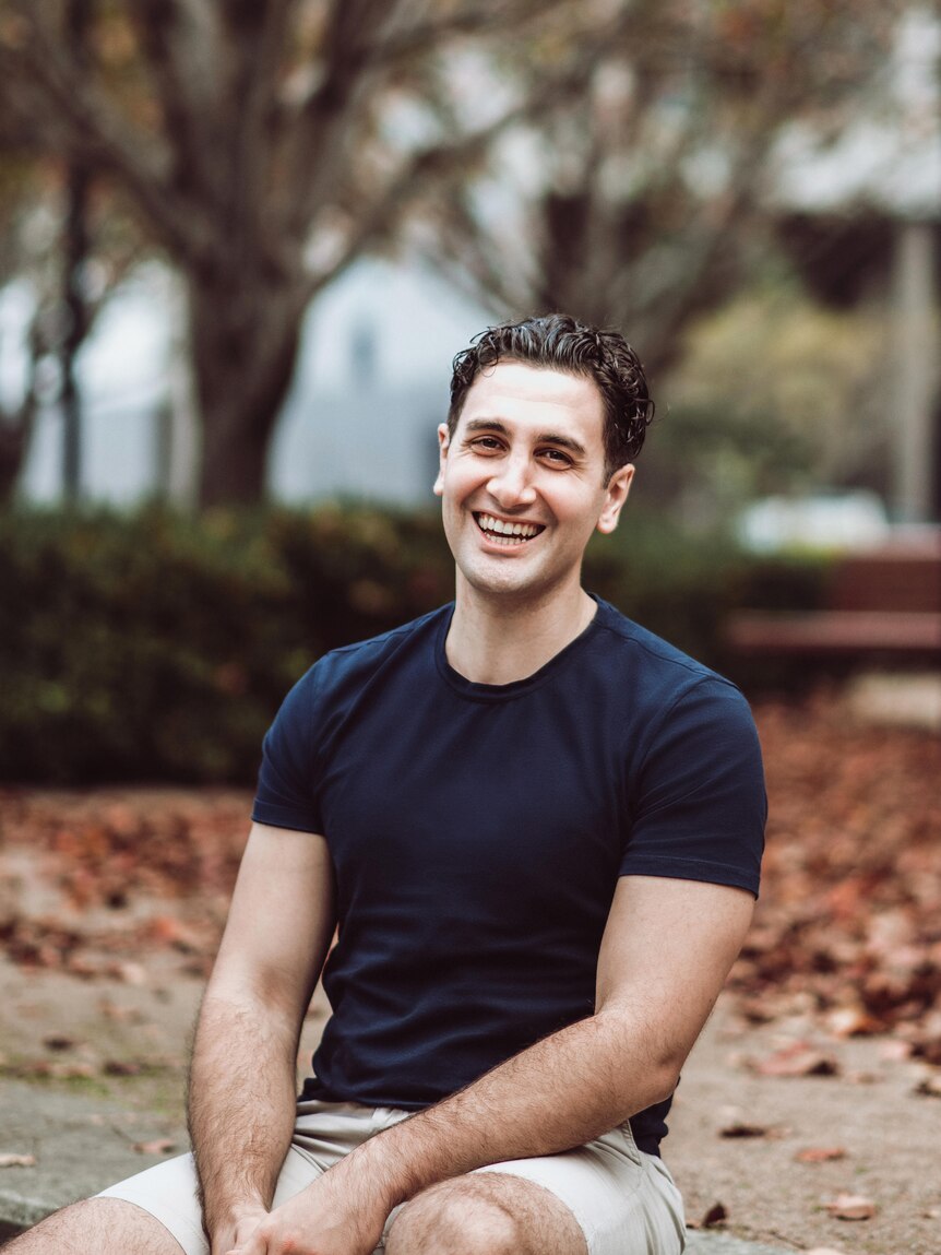 Will Kostakis laughing while seated outside, hands on knees wearing a navy shirt and tan shorts