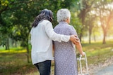 A generic image of a female carer with her arm around an older woman with a walker, as they walk outdoors.