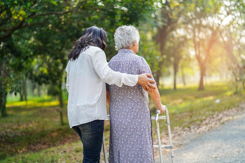 A generic image of a female carer with her arm around an older woman with a walker, as they walk outdoors.