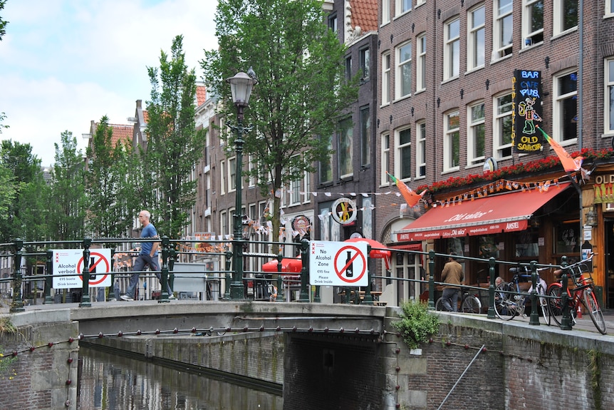 A man is pictured walking over a bridge on a canal in Amsterdam. On the bridge are signs stating that alcohol is not allowed.