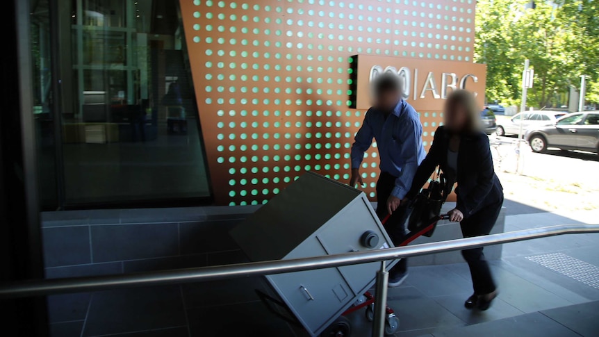 Two people with blurred faces wheel a trolley into the ABC Melbourne office. There is a safe on the trolley.