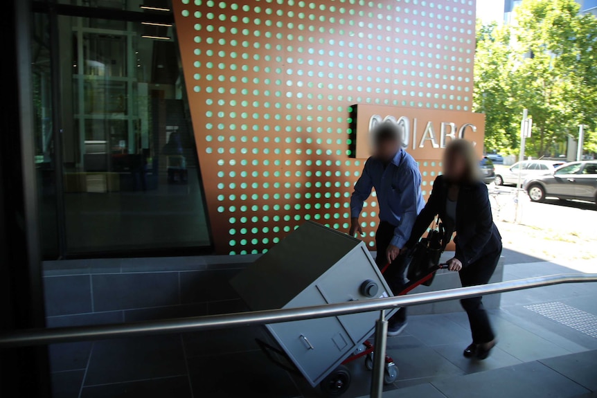 Two people with blurred faces wheel a trolley into the ABC Melbourne office. There is a safe on the trolley.