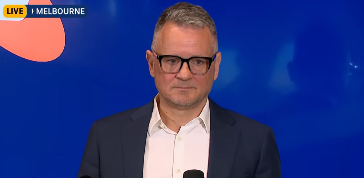 A middle-aged man with glasses speaks into a microphone in front of a blue Telstra backdrop.