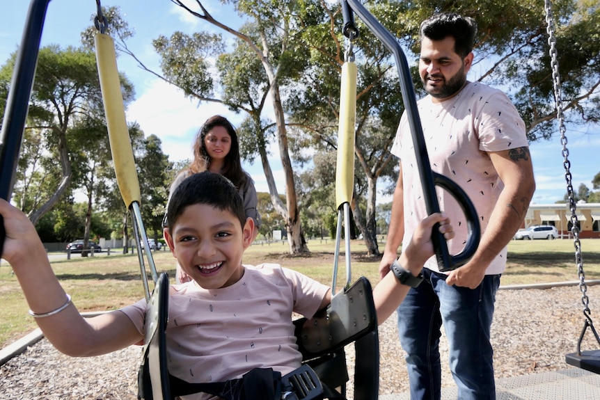At a leafy green park, a young boy is on a swing with a huge smile on his face, while his parents watch on.