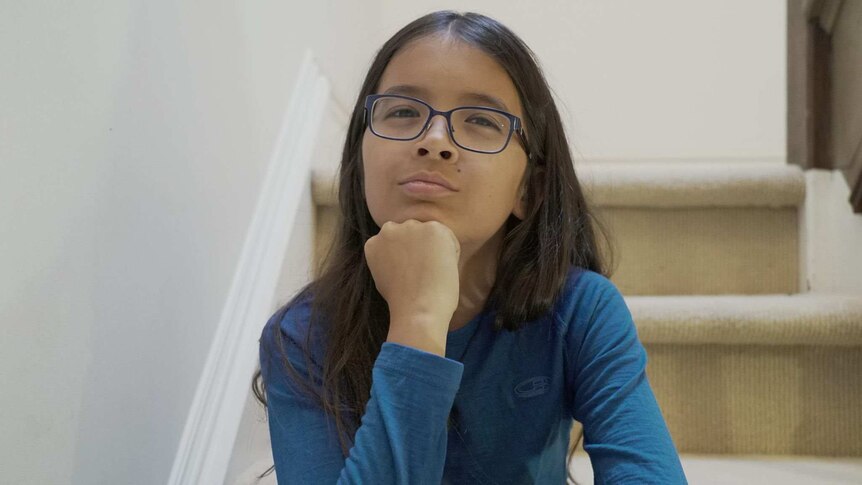 12 year old boy with long brown hair, glasses, blue t-shirt sitting on stairs looking at camera with hand on his chin